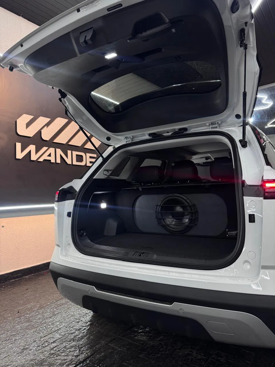 SUV branco com subwoofer de grade no porta-malas, em frente ao letreiro da Wande