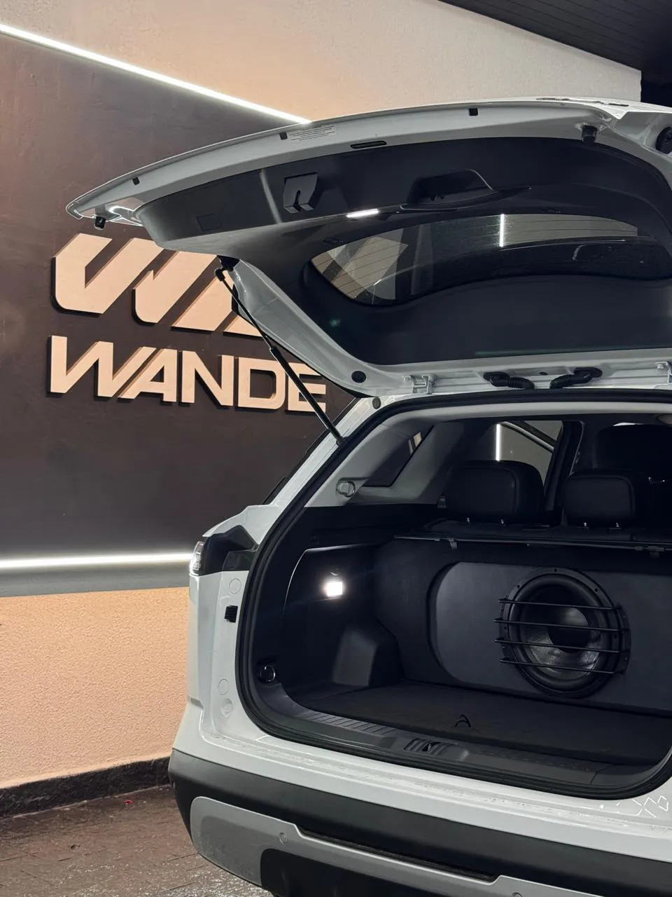 Subwoofer com grade no porta-malas de um SUV branco, com o logo da Wande ao fundo